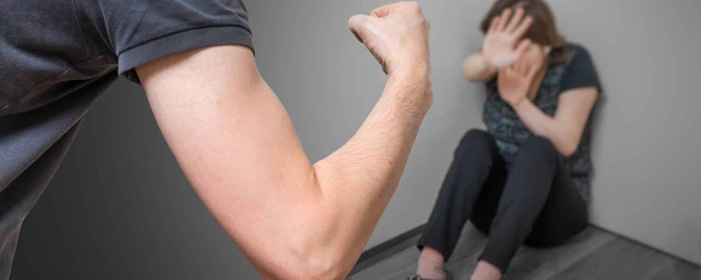 Mann schlägt mit seiner Faust auf einer in der Ecke sitzende, verängstigste Frau.